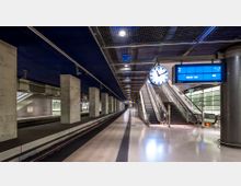 Moderne Bahnhofshalle mit einem überdachten Gleisbereich, hohen Betonpfeilern und einem blauen Deckengitter. Rechts befinden sich eine große Bahnhofsuhr, ein elektronisches Anzeigeboard für Gleis 3 mit der Information „Berlin Hbf“, sowie Rolltreppen und ein Ausfahrtsbereich.