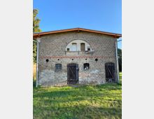 Backsteinbau mit Satteldach in ländlicher Umgebung, ausgestattet mit Holztüren und kleinen Fenstern, teils vergittert; vor dem Gebäude eine Grünfläche und im Hintergrund blauer Himmel.