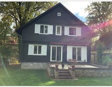 Zweistöckiges schwarzes Haus mit weißen Fensterläden, großen Fenstern und einer Steinterrasse im Vordergrund, umgeben von grünem Rasen und hohen Bäumen. Ein kleiner Weg führt zur Terrasse, die mit Gartenmöbeln und Blumentöpfen dekoriert ist.