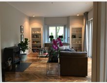 Wohnzimmer mit Holzboden, zentralem Sofa und einer Vase mit violetten Orchideen. Links ein Kaminofen, rechts und mittig Einbauregale mit Dekorationsgegenständen, sowie eine große Fensterfront mit Blick ins Freie.