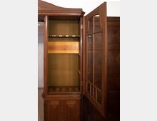 Ein geöffneter, antiker Schrank aus dunkelbraunem Holz mit einer Glastür und sichtbaren Regalfächern. Im Inneren befinden sich vorgesehene Ablagen für Gegenstände sowie Stellflächen mit kreisförmigen Aussparungen, möglicherweise für Gläser oder Flaschen.