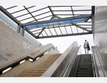Treppenaufgang und Rolltreppe des U-Bahnhofs Rathaus Spandau, überdacht mit einem Glasdach und einer Beschilderung mit der Aufschrift „U-Bhf Rathaus Spandau“. Die Wände sind aus beigem Stein mit Graffiti, und eine Person steht oben an der Rolltreppe.