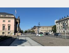 Straßenszene mit historischen Gebäuden in Berlin: Links ein rosafarbenes Gebäude mit klassischen Fenstern und Säulen, rechts weitere helle historische Gebäude. In der Mitte eine gepflasterte Straße mit Autos, Laternen und vereinzeltem Fußgängerverkehr. Im Hintergrund Bäume und ein klarer blauer Himmel.