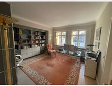 Helles Arbeitszimmer mit einem großen Fenster, das viel Tageslicht hereinlässt. Der Raum enthält eine Wand mit offenen Bücherregalen, einen Glastisch mit Computer, einen Sessel und einen orangen Teppich mit floralem Muster auf Holzboden.