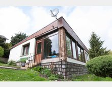 Einstöckiges Wohnhaus mit Flachdach, großen Fenstern und einer Fassade aus weißem Putz und Backstein. Im Vordergrund ist ein leicht geneigter Garten mit Rasen und Sträuchern zu sehen, im Hintergrund stehen Bäume.