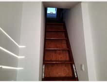 Eine schmale, steile Holztreppe führt durch einen weißen, engen Treppenaufgang nach oben zu einem kleinen Fenster, das Tageslicht hineinlässt. Die Wand links ist von schräg einfallenden Sonnenstrahlen beleuchtet.