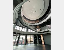 Großer, moderner Innenraum mit einer zentralen, runden Treppenstruktur, die sich spiralförmig nach oben windet. Große Fensterfronten lassen Tageslicht herein, und die Decke ist mit kreisförmigen Elementen gestaltet.