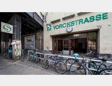 Eingang des S-Bahnhofs Yorckstraße in Berlin mit gelblicher Fassade, einem runden Uhrenschild über der Tür und der grünen Beschriftung "S-Bahn Yorckstraße". Im Vordergrund sind zahlreiche abgestellte Fahrräder und ein Geländer zu sehen, rechts darüber eine Eisenbahnbrücke.