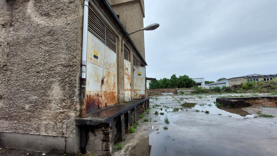 Industriegebäude mit verwitterten, rostigen Metalltoren auf einer erhöhten Rampe. Der umliegende Hof besteht aus Betonflächen mit vereinzeltem Grasbewuchs und Pfützen, im Hintergrund sind weitere Gebäude und Bäume zu sehen.