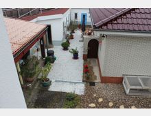 Ein Innenhof mit Kiesboden, umgeben von Gebäuden mit roten Ziegeldächern und weiß gestrichenen Wänden. Der Hof ist dekoriert mit Blumentöpfen, bunten Blumen und Sitzmöbeln; im Hintergrund ist eine blaue Tür zu sehen.