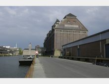 Industriehafen mit großen Backsteingebäuden, darunter ein als „Getreidespeicher“ beschriftetes Gebäude. Im Vordergrund ein Wasserweg mit Anlegestellen und eine parallel verlaufende Straße, im Hintergrund weitere Industriegebäude und ein Turm.