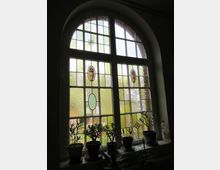 Großes, bogenförmiges Fenster mit farbigem Buntglas und dekorativen Mustern, darunter ovale und wappenähnliche Ornamente. Auf der Fensterbank stehen mehrere Topfpflanzen, während im Hintergrund ein Ausblick auf Bäume und Gebäude erkennbar ist.