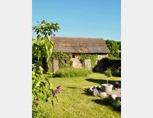 Ein alter, mit Efeu bewachsener Schuppen aus Ziegelsteinen mit Holztüren steht in einer grünen Gartenlandschaft. Der Vordergrund zeigt eine Rasenfläche mit Blühpflanzen, einem Pflanzenkübel und dekorativen Steinen.