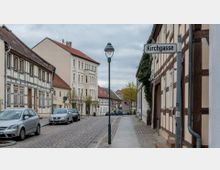 Straßenszene einer kleinen Altstadt mit gepflasterter Fahrbahn, links geparkten Autos und Fachwerkhäusern auf beiden Seiten. Ein Straßenschild mit der Aufschrift „Kirchgasse“ ist rechts sichtbar, und im Hintergrund befinden sich weitere historische Gebäude.