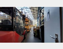 Innenansicht eines großen Lagers mit hohen Regalen, die mit Waren bestückt sind, und mehreren Gabelstaplern in den Gängen. Die Decke besteht aus einem Stahlgerüst mit Oberlichtern, die Tageslicht ins Gebäude lassen.