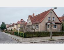 Straßenszene mit Einfamilienhäusern im Altbaustil, erkennbar an den verputzten Fassaden und Satteldächern mit roten Ziegeln. Im Vordergrund sind ein Gehweg, ein Straßenschild und eine Bushaltestelle zu sehen, umgeben von niedrigen Hecken und kahlen Bäumen.