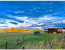 Eine kleine Flugzeug-Landebahn auf einer Wiese mit einem blauen Leichtflugzeug, einem hölzernen Gebäude und einem roten Versorgungsfahrzeug. Im Hintergrund Herbstbäume und ein weitläufiger Himmel mit Wolkenformationen.