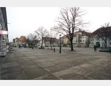 Weitläufiger, gepflasterter Marktplatz mit vereinzelt stehenden Bäumen, umgeben von historischen Gebäuden mit roten Dächern und verzierten Fassaden. Im Hintergrund und an den Seiten verlaufen Gehwege und kleinere Geschäfte.