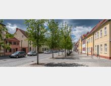 Straßenszene in einer Kleinstadt mit gepflastertem Bürgersteig, Bäumen in einer Reihe und zweigeschossigen Häusern mit farbenfrohen Fassaden und Satteldächern. Autos parken auf der gepflasterten Straße, während im Hintergrund Wolken am Himmel zu sehen sind.