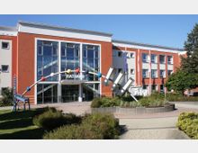 Modernes Rathausgebäude mit rot-weißer Fassade und großen Glasfenstern im Eingangsbereich. Davor steht eine bunte Kunstinstallation mit Kugeln und Metallstäben, umgeben von Pflanzbeeten und Gehwegen.