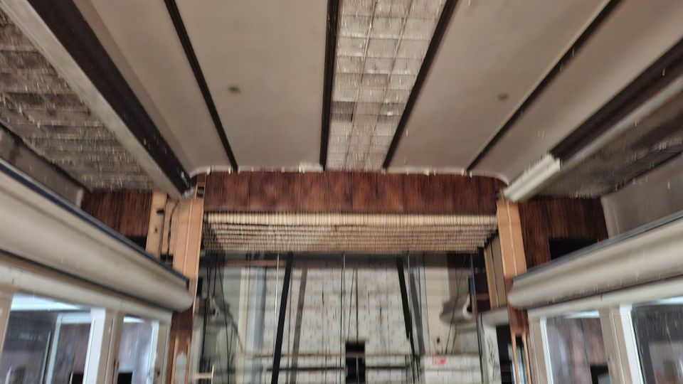 Blick nach oben in einen Industrie- bzw. Werkstattraum mit breiten Deckenbalken, großen Raster-Leuchtpanelen und freigelegten Holz- und Metallkonstruktionen. Seitlich sind Fensterfronten und Stahlträger sichtbar, im Hintergrund eine geflieste Wand mit Kabeln und Installationen.