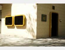 Außenansicht eines Gebäudes mit beigefarbenen Fliesenwänden, gelben, rechteckigen Schaukästen an der Wand und einer dunklen Eingangstür mit goldenem Rahmen. Daneben befindet sich ein Schild mit der Aufschrift „Rum Trader“.