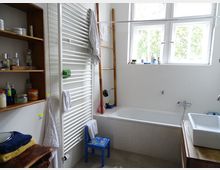 Ein Badezimmer mit weiß gefliesten Wänden, einer Badewanne, einem rechteckigen weißen Waschbecken auf einem Holzschrank, einem Heizkörper-Handtuchhalter und einem Holzregal mit Pflegeprodukten. Ein blauer Kinderstuhl steht neben der Badewanne, und ein Fenster mit Blick auf grüne Bäume lässt Tageslicht herein.