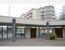 Eingangsbereich mit Tor und Überdachung aus Beton, im Hintergrund ein mehrstöckiges Wohngebäude mit Balkonen und großen Fenstern. Der Vorplatz ist mit Pflastersteinen ausgelegt, daneben steht ein kleiner Strauch in einem Pflanzkübel.