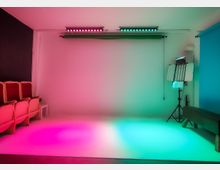 Ein Fotostudio mit einem weißen Hintergrund, dynamisch beleuchtet in rosa und grün. Links befinden sich rote Kinosessel, rechts ein Lichtstativ und eine blaue Stoffrolle.