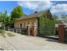 Ein kleiner, historischer Gebäudekomplex mit gelb gestrichener Fassade und klassizistischen Verzierungen, umgeben von einer Zaunanlage mit rotem Ziegelstein und schwarzen Metalltoren. Eine Seite des Hauses ist dicht mit Efeu bewachsen; im Hintergrund sind Bäume und ein blauer Himmel mit vereinzelten Wolken zu sehen.