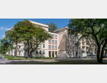 Mehrstöckiges Wohngebäude im Stil der Nachkriegsmoderne mit beigen Fassaden und hohen Fenstern, umgeben von Bäumen und begrünten Flächen; im Vordergrund eine Straße und geparkte Autos.