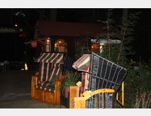 Ein Außenbereich mit mehreren Strandkörben vor einem kleinen Gebäude mit rotbraunem Dach und erleuchteten Fenstern. Im Hintergrund sind Pflanzen und ein Schild mit der Aufschrift "Waldhütte" sichtbar.