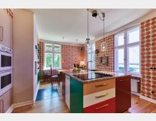 Bunte Küche mit einer zentralen Kochinsel, die Schubladen in verschiedenen Farben hat, einer Granit-Arbeitsplatte und hängenden Glaslampen. Die Wände sind mit gemusterten Tapeten dekoriert, und große Fenster lassen viel Tageslicht herein.