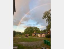 Ein ländlicher Hof mit einer kleinen Backsteinhütte und Nebengebäuden, umgeben von viel Grün und Bäumen. Ein Regenbogen erstreckt sich über den Himmel, während Pflastersteine, eine Wiese und grüne Mülltonnen im Vordergrund sichtbar sind.