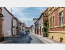 Eine schmale, gepflasterte Straße in einer Altstadt, gesäumt von Fachwerkhäusern und Gebäuden mit Backsteinfassaden. Geparkte Autos befinden sich entlang der Straße, im Hintergrund sind weitere Häuser und ein blauer Himmel zu sehen.