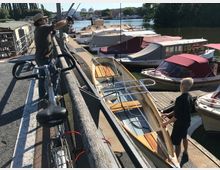 Zwei Personen an einem Bootssteg neben mehreren vertäuten, überdachten Motorbooten. Im Vordergrund ein Fahrrad, im Hintergrund eine Flusslandschaft mit Bäumen und Gebäuden.