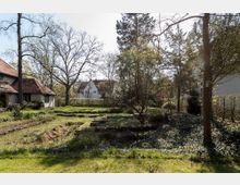 Gartenlandschaft mit Rasenflächen, vereinzelten Bäumen und kleinen Sträuchern im Vordergrund, umgeben von Wohnhäusern mit roten Ziegeldächern. Im Hintergrund stehen weitere Häuser sowie dichte Hecken, die den Bereich abgrenzen.