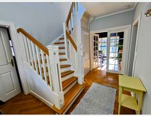 Ein Eingangsbereich in einem hellen Wohnhaus mit einer weißen Holztreppe mit Holzstufen, die nach oben führt. Der Boden besteht aus Fischgrätparkett, und im Hintergrund ist ein Raum mit bodentiefen Glastüren sichtbar, durch die viel Tageslicht einfällt.