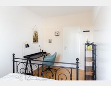 Helles Schlafzimmer mit einem schwarzen Metallbettgestell im Vordergrund, einem schwarzen Schreibtisch mit Lampe und einem hellblauen Stuhl. An der Wand hängt ein Pflanzenposter sowie kleine Wandregale, daneben ein offenes Regal mit Deko und Pflanzen.