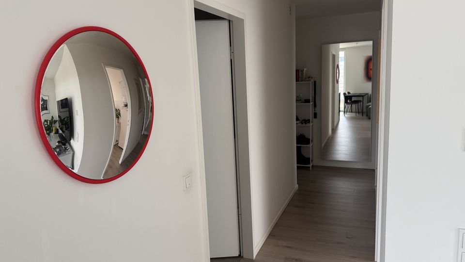 Heller Wohnungsflur mit weißen Wänden und dunkelgrauem Holzfußboden; links hängt ein runder Spiegel mit rotem Rahmen an der Wand. Am Ende des Gangs führt eine offene Tür in einen weiteren Raum, daneben steht ein schmales Regal.