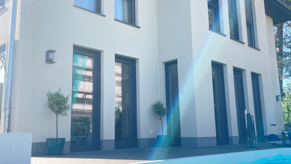 Moderne zweistöckige Villa mit weißer Fassade und großen, dunklen Fenstern, umgeben von einer Terrasse. Im Vordergrund ein Swimmingpool mit klarem, blauem Wasser; Bäume sind im Hintergrund sichtbar.