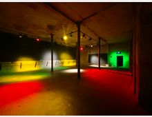 Ein leerer Raum in einem industriellen Stil mit Betonwänden, Säulen und einer hohen Decke. Farbige Beleuchtung in Rot, Grün und Gelb erhellt den Boden und die Wände, während ein Ausgang mit grün beleuchtetem Rahmen im Hintergrund sichtbar ist.