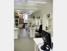 Modernes Büro mit weißen Möbeln und offenen Regalen, mehreren Schreibtischen und Stühlen. Im Hintergrund führt eine weiße Tür mit Glasfenster nach draußen, umgeben von einigen Stufen und Bildern an den Wänden.