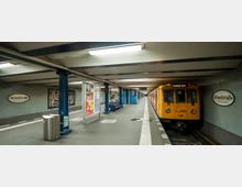U-Bahnhof mit gelbem Zug an einer Bahnsteigkante, beleuchtet durch Deckenleuchten. Die Wände sind mit hellblauen Fliesen verkleidet, der Bahnsteig trägt das Stationsschild „Uhlandstraße“.