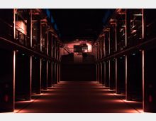 Industriehalle mit symmetrisch angeordneten Säulenreihen, die von orangefarbenem Licht beleuchtet werden. Im Hintergrund befindet sich eine erhöhte Plattform mit Treppen und Industriegeräten.