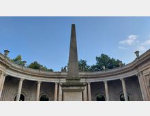 Steinerner Obelisk in der Mitte eines offenen, halbkreisförmigen Kolonnadenbaus mit Säulen und Reliefs, umgeben von Bäumen und unter einem klaren Himmel.