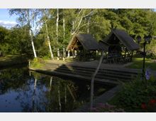 Zwei überdachte Sitzbereiche aus Holz mit Strohdächern stehen auf einer Plattform neben einem ruhigen Gewässer, umgeben von Bäumen, Wiese und Blumenbeeten. Im Vordergrund sind ein Holzsteg und Laternen sichtbar, die Szene erinnert an einen naturnahen Park oder Rastplatz.