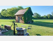 Kleines Backsteinhaus mit Dach aus roten Ziegeln, teilweise mit Efeu bewachsen, auf einer großen Grasfläche. Im Vordergrund stehen ein alter Brunnen mit Sitzmöbeln aus Holz und Metall sowie eine rustikale Gartenbank, umgeben von Bäumen und einem weiten Feld im Hintergrund.