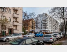 Straßenszene in einem Wohngebiet mit mehrstöckigen Wohnhäusern im Hintergrund, darunter ein modernes weißes Gebäude mit Balkonen und ein älteres Gebäude mit roten Ziegelkanten. Im Vordergrund parken mehrere Autos entlang einer gepflasterten Straße, umgeben von kahlen Bäumen.