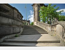 Steintreppe mit verzierten Geländern und einer hohen Steinmauer, die zu einem historischen Torbogen führt, umgeben von Bäumen und einem klaren Himmel. Im Hintergrund sind weitere architektonische Elemente wie Balustraden und Säulen zu sehen.